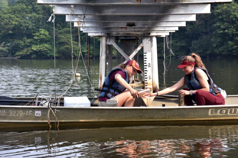 Two women collect water samples on a boat under a bridge. 